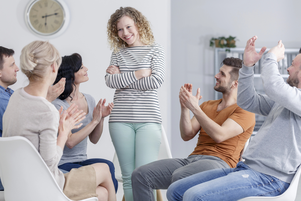 A woman stands and speaks while others clap in an intensive outpatient program group session